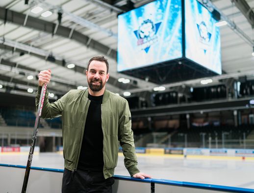 Derek Dinger hält einen Eishockeyschläger in der Hand und lehnt an der Bande. 
