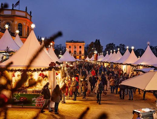 Sie sehen die aufgebauten Stände vor der Orangerie. Menschen laufen zwischen den weihnachtlichen Ständen her. 